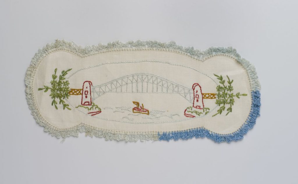 A hand embroidered cream linen sandwich doily. It is rectangular in shape and bulges at both ends. It features a Sydney Harbour Bridge design in red, light blue, green and brown thread. On the water underneath the bridge is a small boat, embroidered in red and brown thread. The bridge and boat are encircled by an oval stitched with light blue thread and there is a bouquet of green leaves at each end. The edge has been crocheted in two different shades of blue.